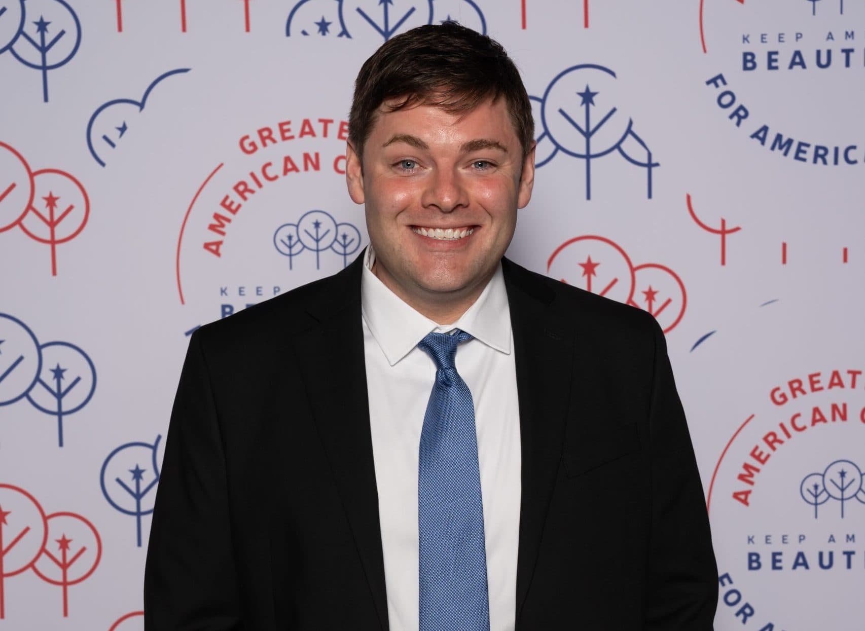 Solo portrait of a young man in a black suit, white shirt and light blue tie against the tree-patterned wall
