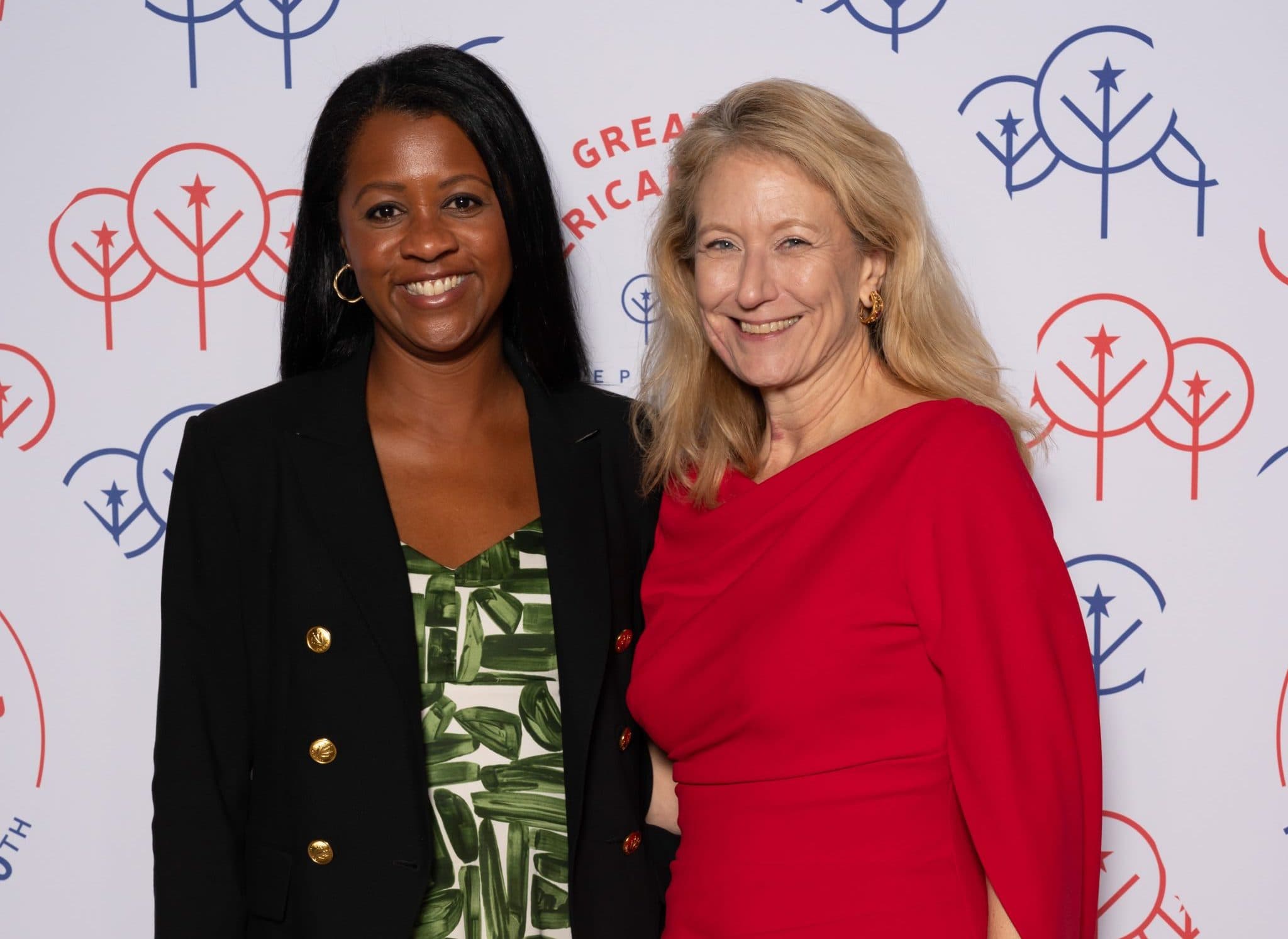 Woman in a black blazer with gold buttons over a green brushstroke-print dress standing with a blonde woman in a red draped dress