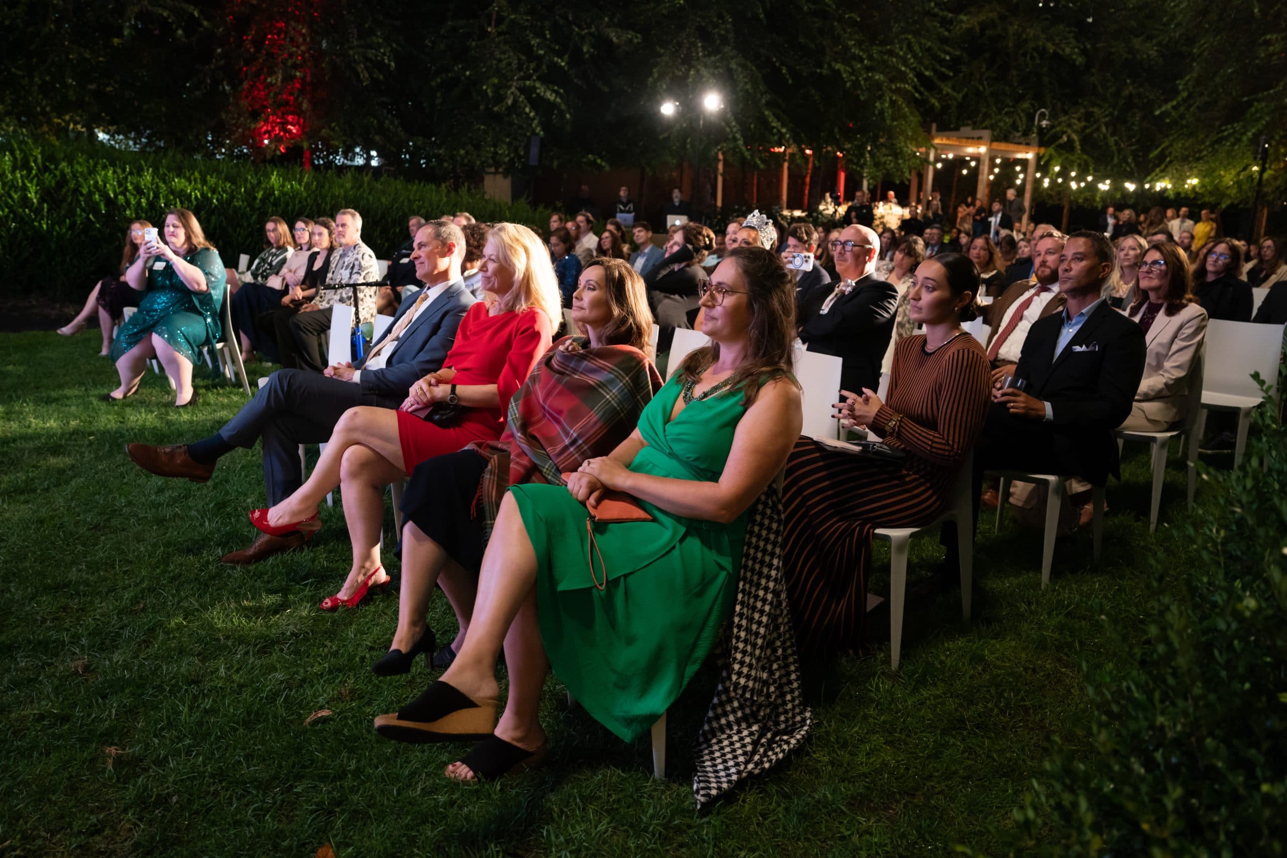 Seated audience on white chairs on a lawn at night, front row including a woman in red and a woman in a green dress with a plaid wrap
