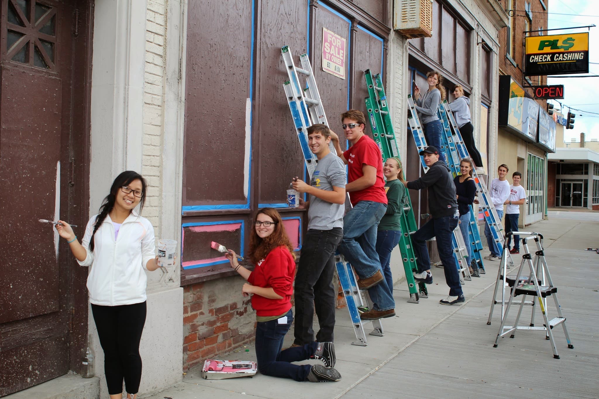 A dozen volunteers with brushes and ladders repaint a boarded-up downtown storefront; a PLS Check Cashing sign and a For Sale By Owner notice hang nearby