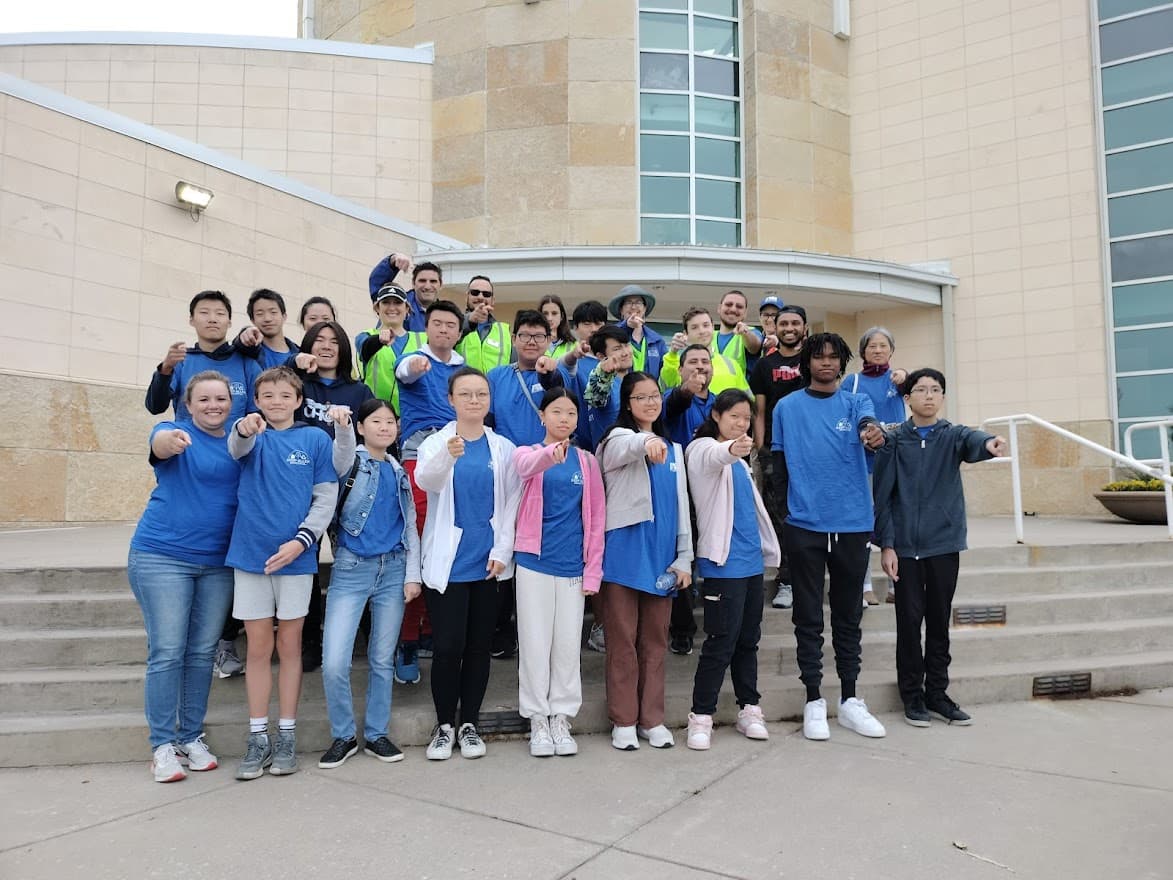 Small group photo of roughly twenty students in blue T-shirts pointing at the camera on the steps of a stone civic building