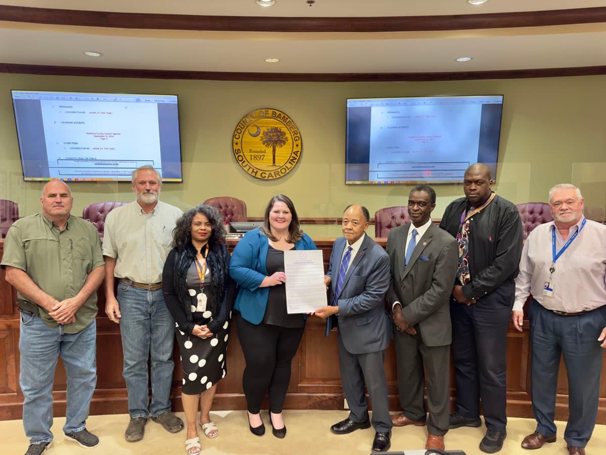 Nine officials line up in a county council chamber, two of them holding a signed declaration beneath the County of Bamberg, South Carolina seal