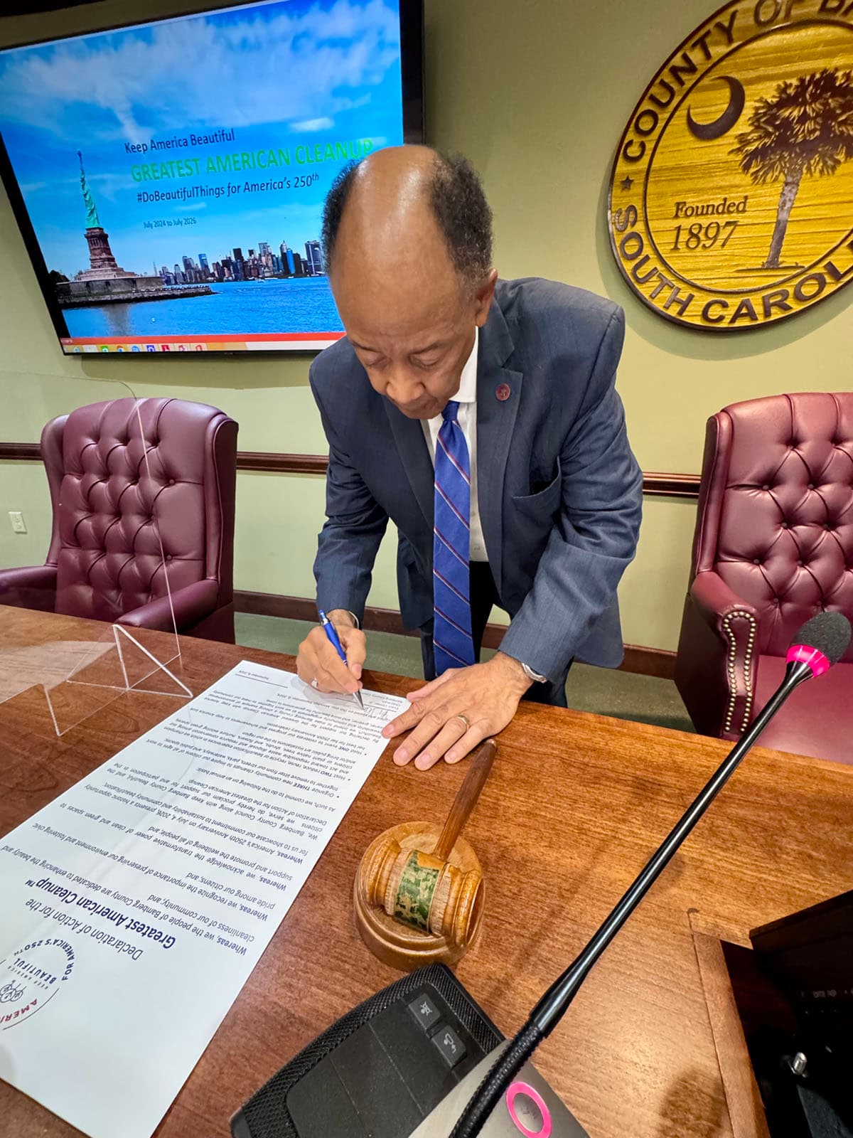 A man in a grey suit signs the Greatest American Cleanup Declaration of Action at a council table beside a gavel; the screen behind shows a Statue of Liberty campaign slide