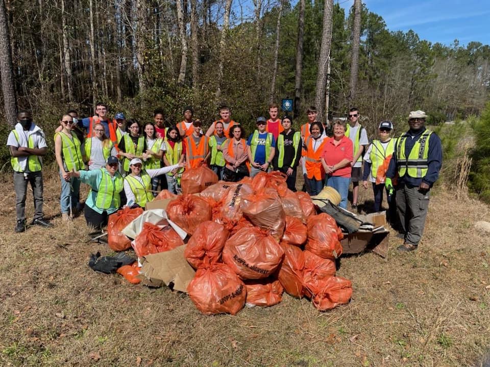 About thirty volunteers in orange and lime safety vests stand behind a mound of orange trash bags at a pine woodland roadside