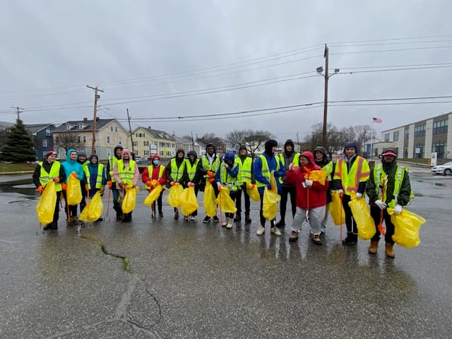 A long line of volunteers in raincoats and hi-vis vests hold yellow trash bags on a wet parking lot under grey skies, houses and a US flag behind