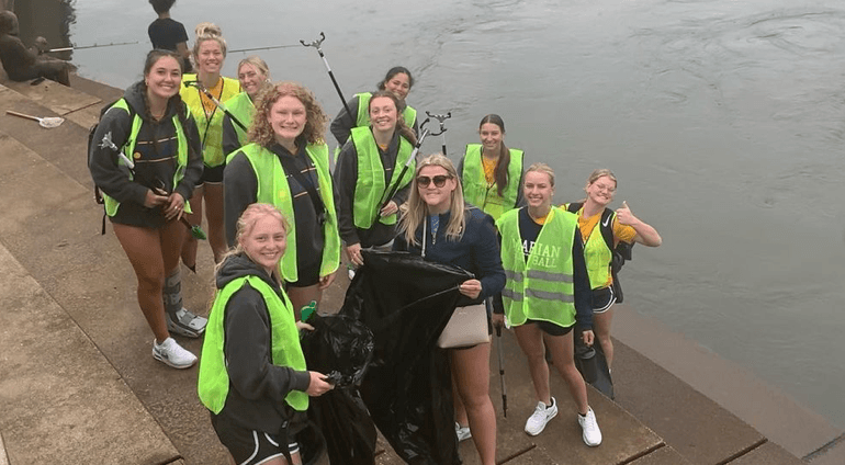 Cropped thumbnail of a dozen young women in neon vests holding grabbers and a black bag on stone steps beside a river