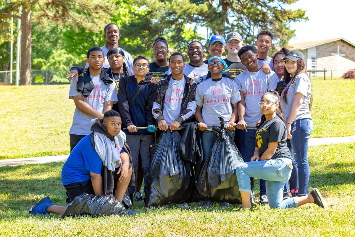 About seventeen high-school students in grey Keep America Beautiful shirts kneel and stand around two full black trash bags on a sunlit lawn