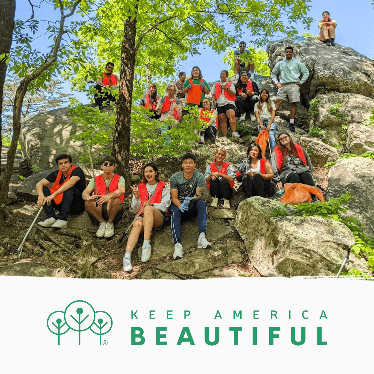 Twenty volunteers in orange vests sit across mossy boulders in a green forest, with the Keep America Beautiful tree logo on a white band below