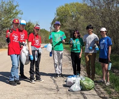 Seven teenagers with gloves and trash bags stand on a rural gravel roadside, three of them in red club shirts, bagged litter at their feet