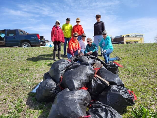 Eight volunteers in caps stand on a grassy slope behind a long row of black trash bags; a pickup truck and a McDonald's sign in the background