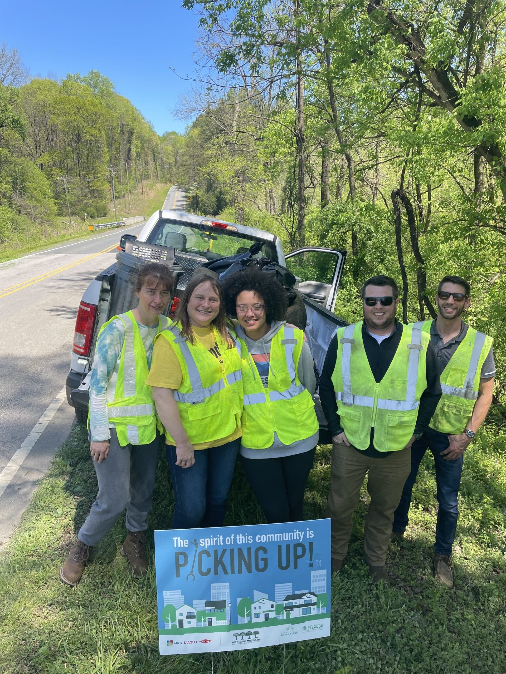 Five volunteers in lime safety vests stand at a pickup truck on a wooded two-lane road behind a yard sign reading The spirit of this community is PICKING UP!