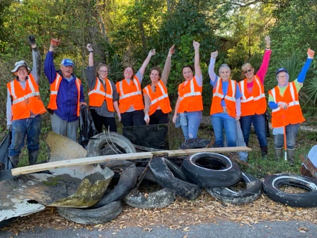 Ten volunteers in orange vests raise their fists behind a roadside heap of scrap tyres, timber and torn metal they have hauled out