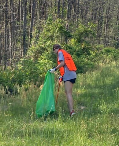 A lone volunteer in an orange vest drags a green bag through tall roadside grass at the edge of a pine wood