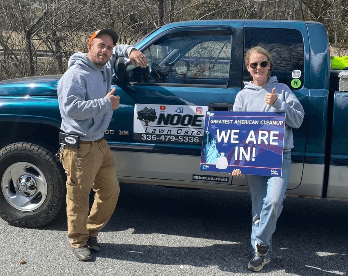 A man and a woman give thumbs up beside a teal Nooe Lawn Care pickup; she holds a Greatest American Cleanup WE ARE IN! yard sign