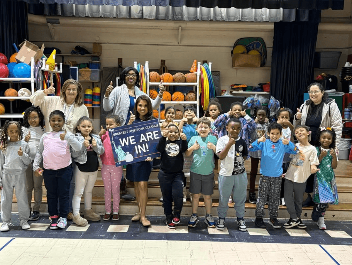 A class of young children and four adults give thumbs up in a school gym, a teacher holding a Greatest American Cleanup WE ARE IN! sign