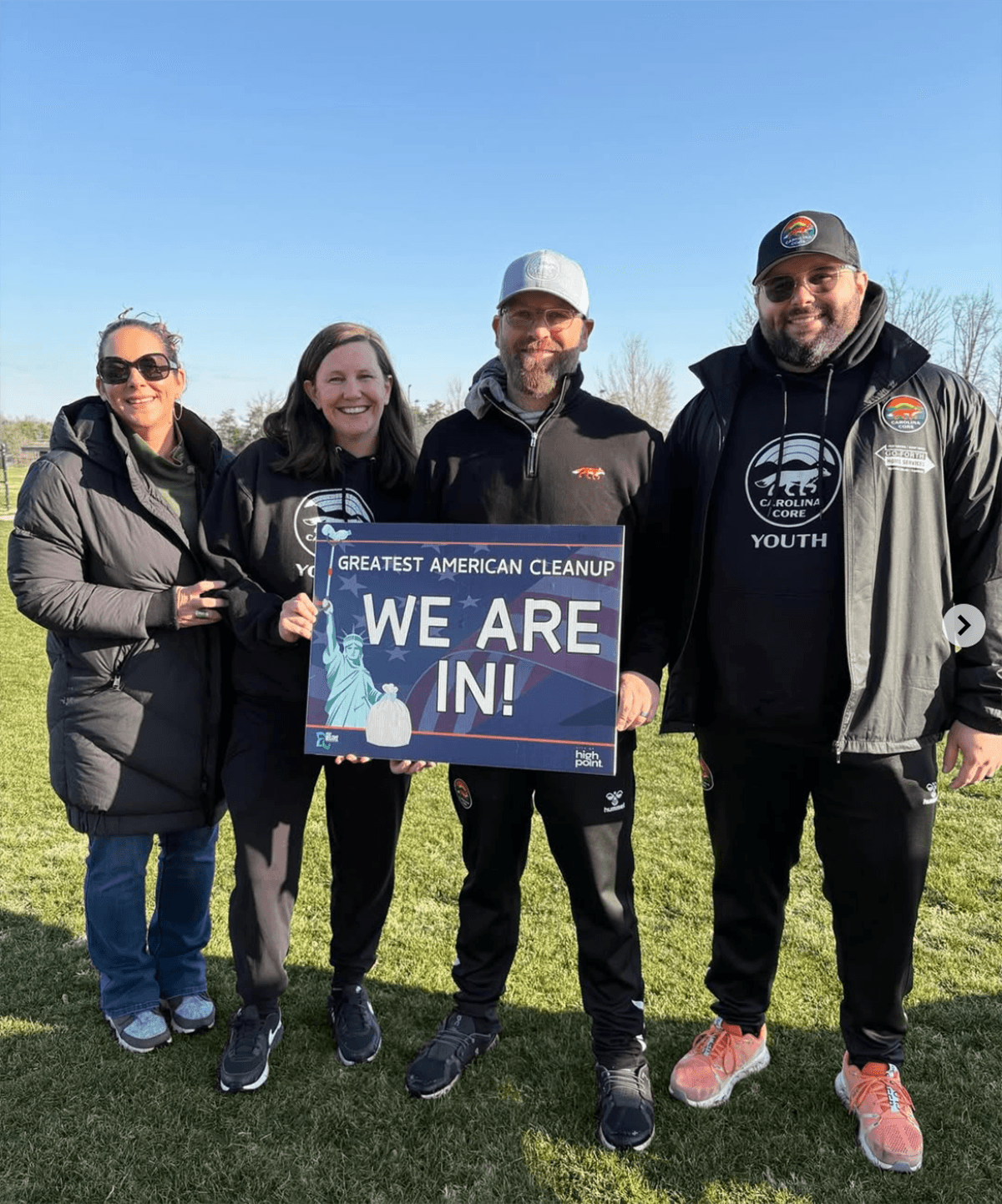 Four adults in black Carolina Core Youth jackets stand on a winter field holding a Greatest American Cleanup WE ARE IN! sign