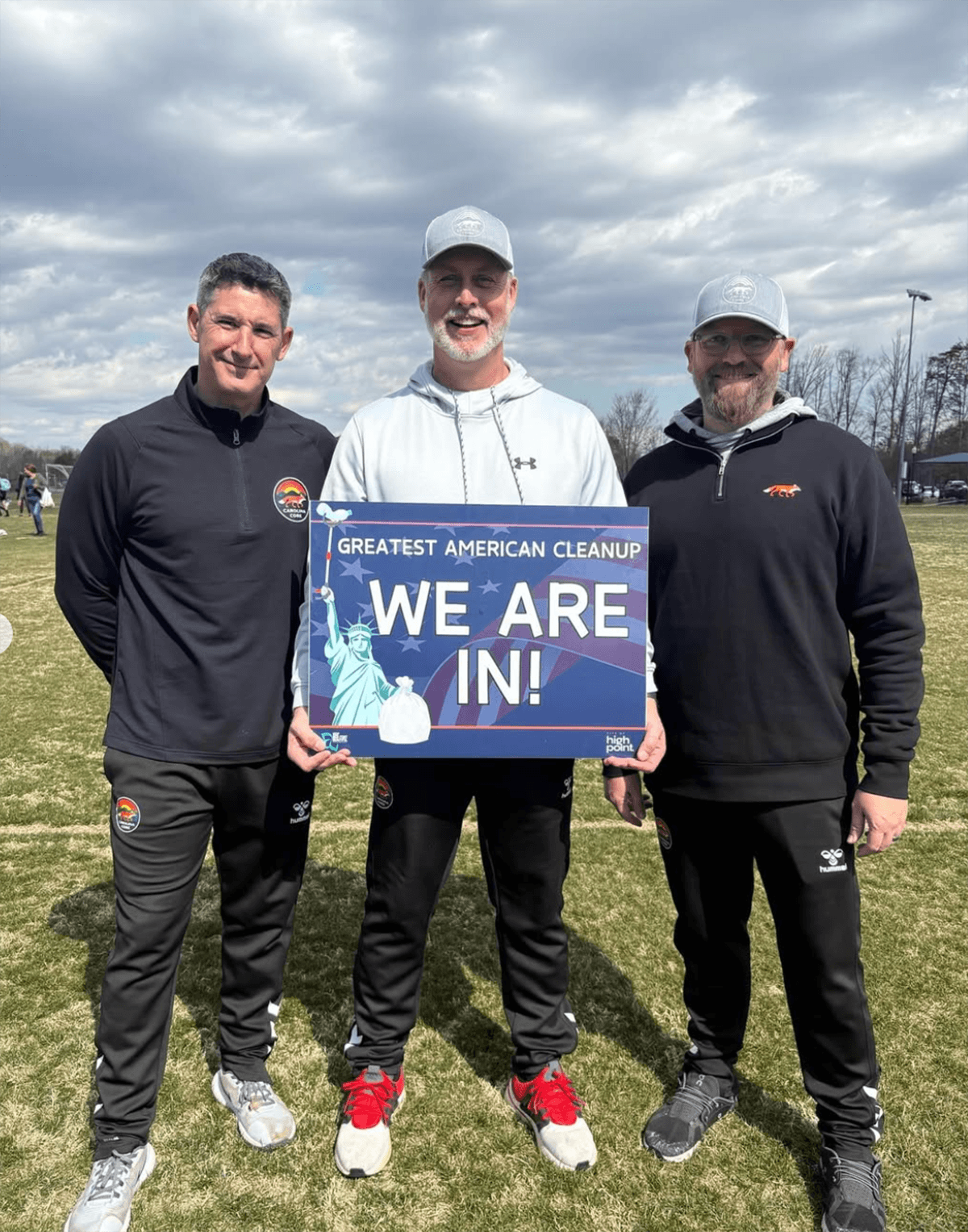 Three men in soccer coaching gear stand on a grass pitch under cloudy skies with a Greatest American Cleanup WE ARE IN! sign between them
