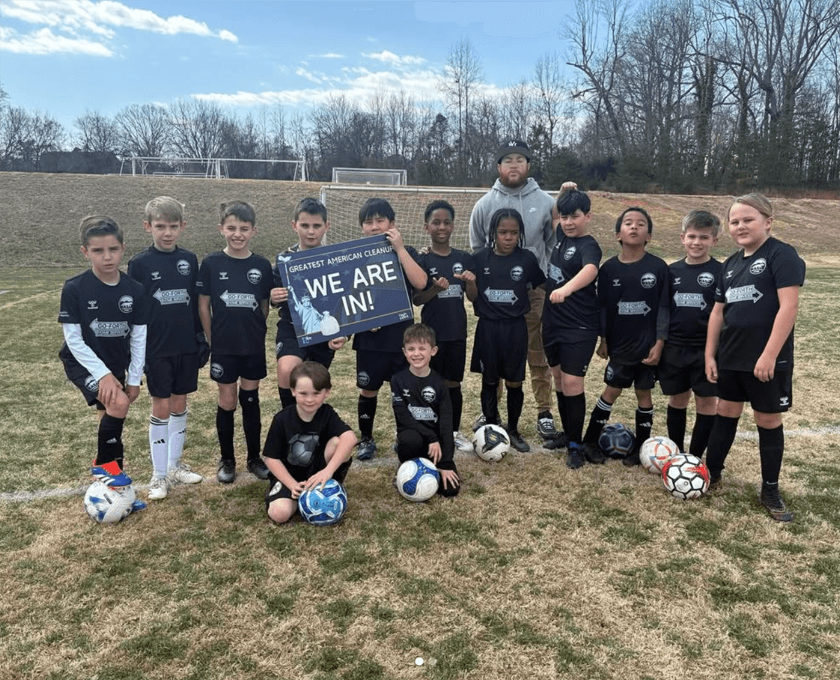 A boys' youth soccer team in black kit and their coach pose with footballs and a Greatest American Cleanup WE ARE IN! sign