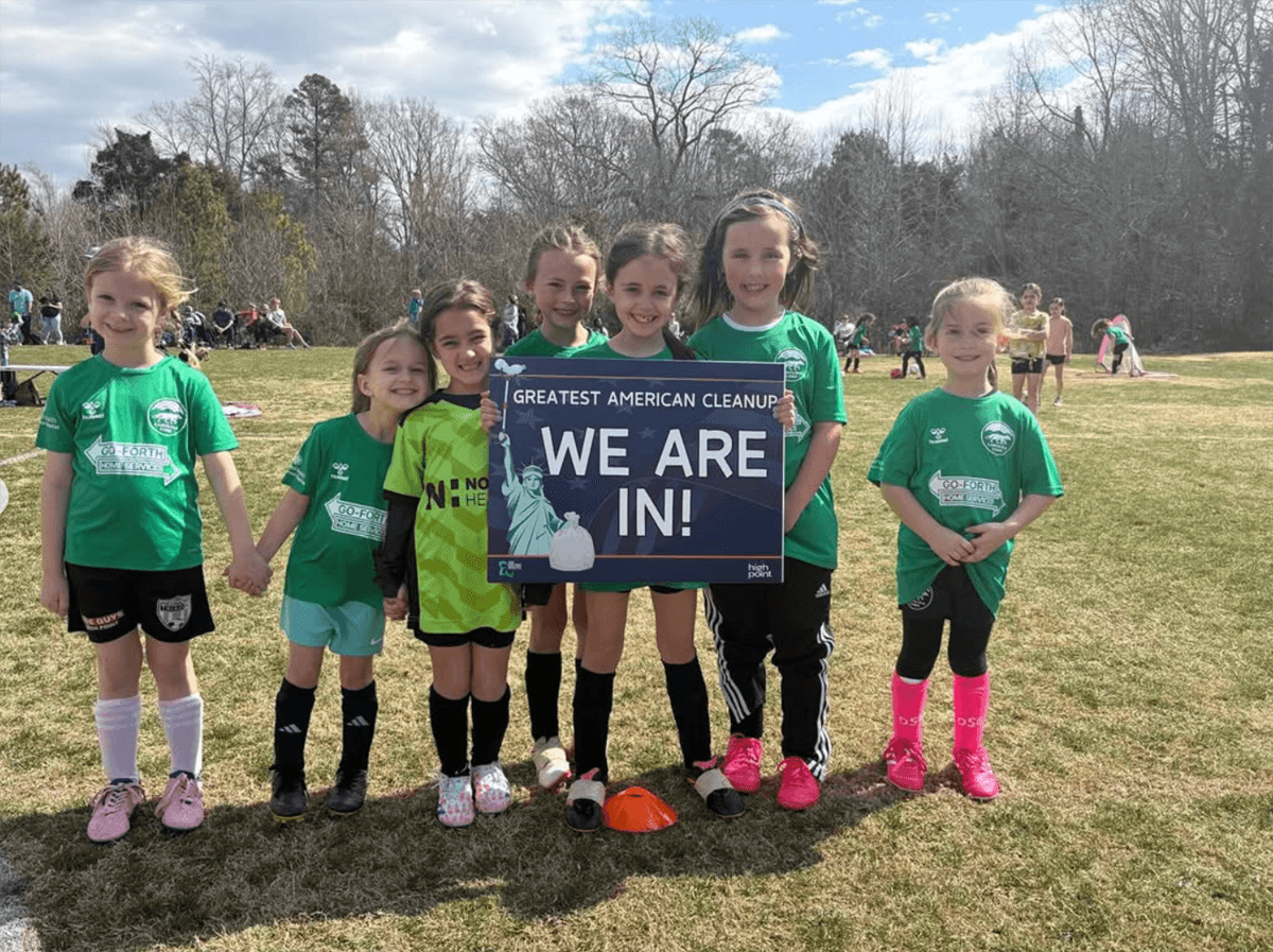Seven girls in green soccer shirts hold a Greatest American Cleanup WE ARE IN! sign on a winter playing field
