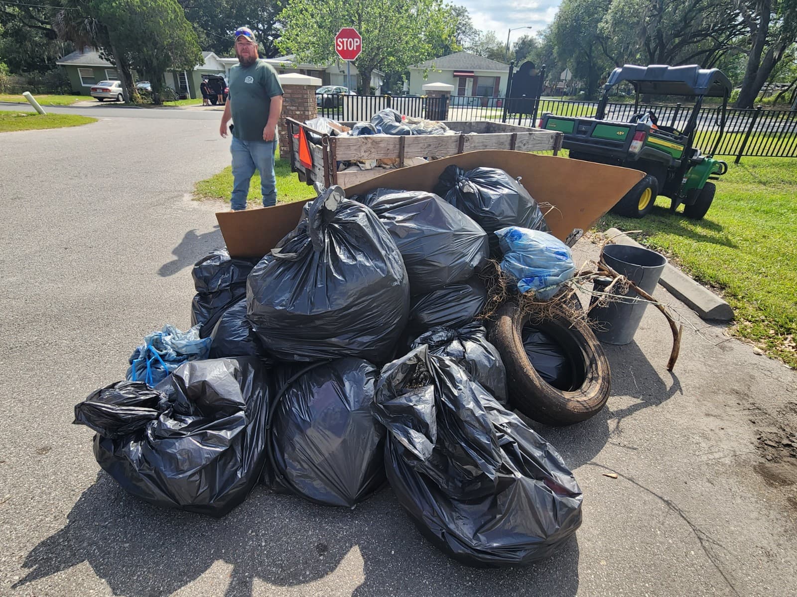 A bearded man in a green shirt stands by a street-corner pile of black bags, an old tyre and a loaded utility trailer with a John Deere Gator behind
