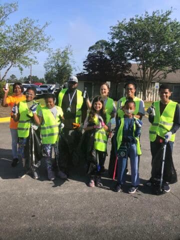 Ten adults and children in lime hi-vis vests hold trash bags and grabbers in a parking lot outside a low brick building