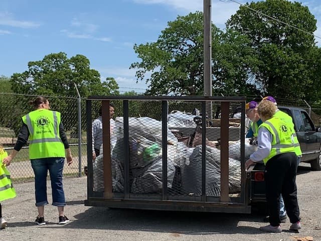 Lions Club volunteers in lime vests load a caged trailer packed with bagged litter on a gravel lot behind a chain-link fence