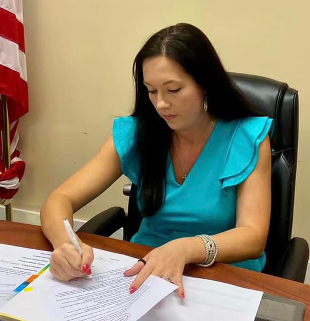 A dark-haired woman in a teal blouse signs a document at an office desk, a US flag standing behind her