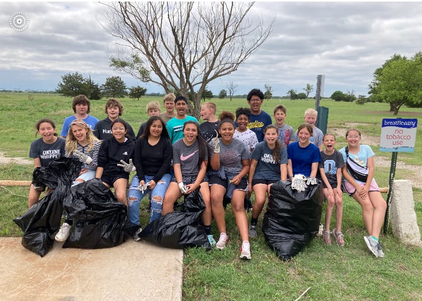 Thumbnail of about twenty middle-school students seated in a field with black bags at their feet under a cloudy prairie sky