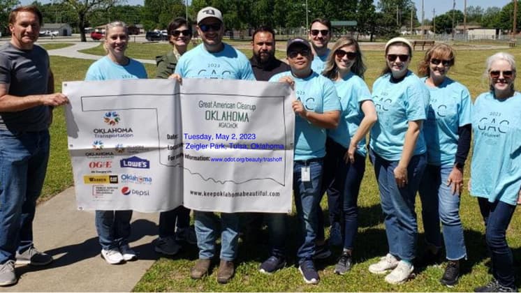 Small photo of a dozen people in pale blue shirts holding a Great American Cleanup Oklahoma banner on a park lawn
