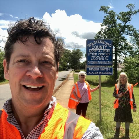 Selfie of a man in an orange safety vest with two women holding grabbers behind him at a blue Spartanburg County Adopt-A-Road Litter Control Program sign