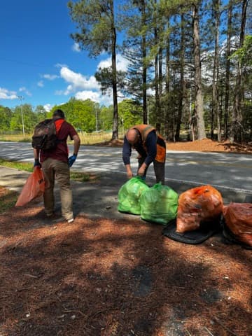 Two men bag litter on a pine-needle roadside, green and orange collection bags standing on the ground beside them