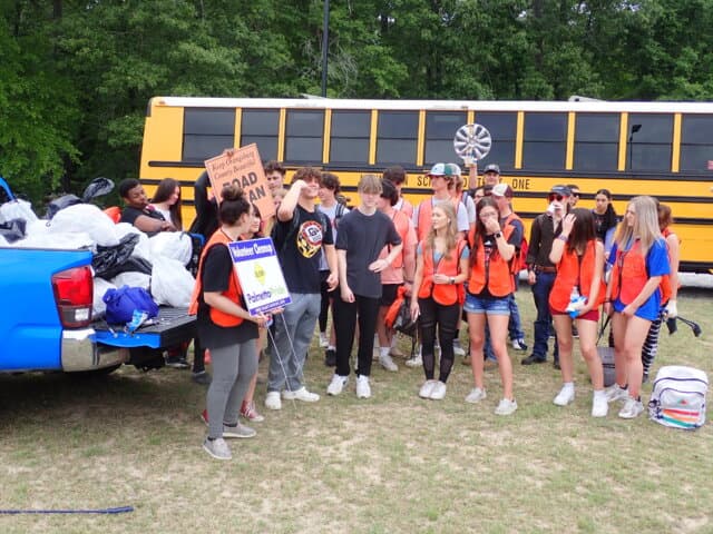 Teenagers in orange vests gather between a yellow school bus and a blue pickup piled with collected bags; a volunteer holds a Road Ban cleanup sign