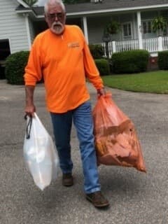 An older man in an orange long-sleeved shirt carries a white bag and a full orange litter bag up a residential driveway