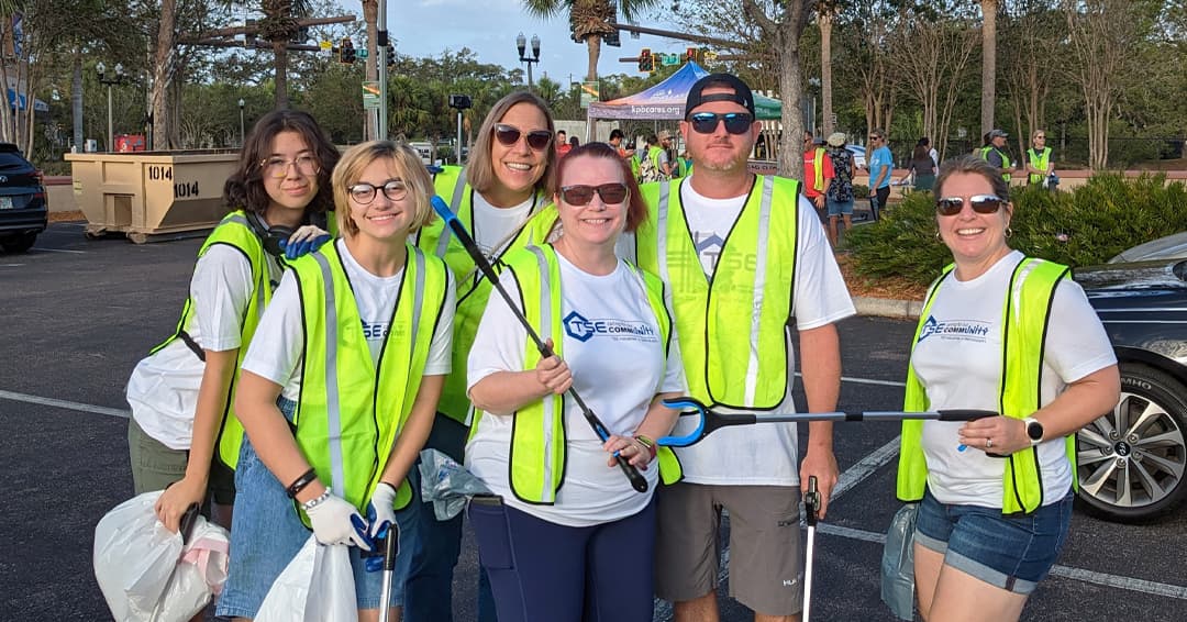 Wide thumbnail of six volunteers in yellow vests and matching white team shirts crowded together with litter grabbers in a sunlit parking lot
