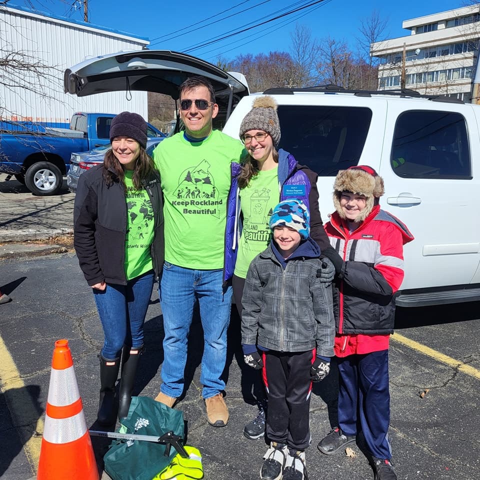 Square crop of a family of five in winter coats posing behind an open SUV hatch next to an orange traffic cone