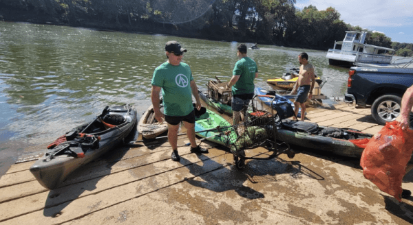 Men in green shirts unload kayaks loaded with hauled-out debris onto a wooden boat ramp at a river, a red trash bag at the edge of the frame