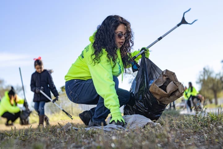 Woman in a neon hoodie and sunglasses crouches in dry grass filling a black bag with a grabber, a child and other volunteers blurred behind her
