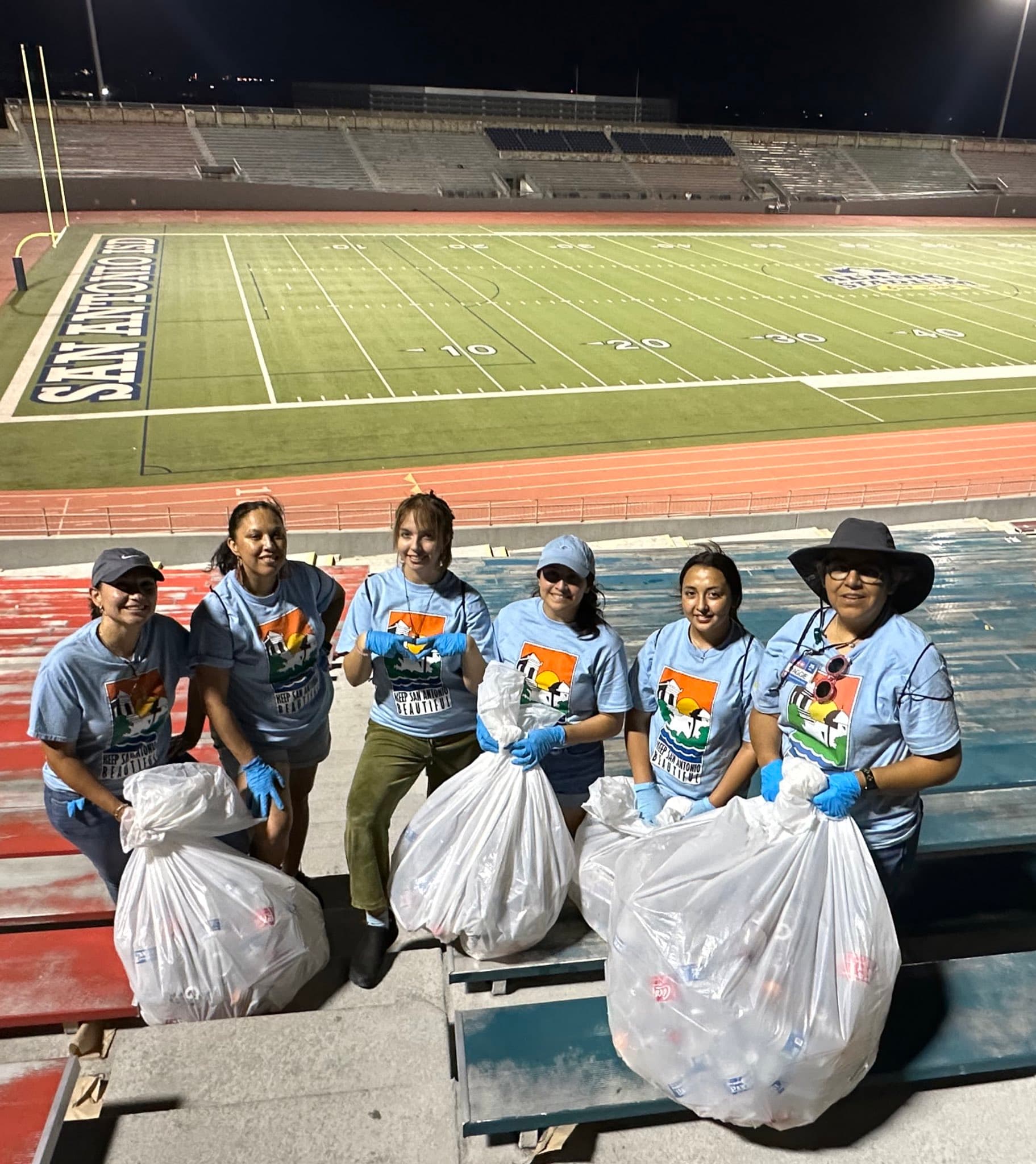 Six volunteers in "Keep San Antonio Beautiful" shirts hold white bags of collected bottles on stadium bleachers at night, the San Antonio ISD field lit behind them