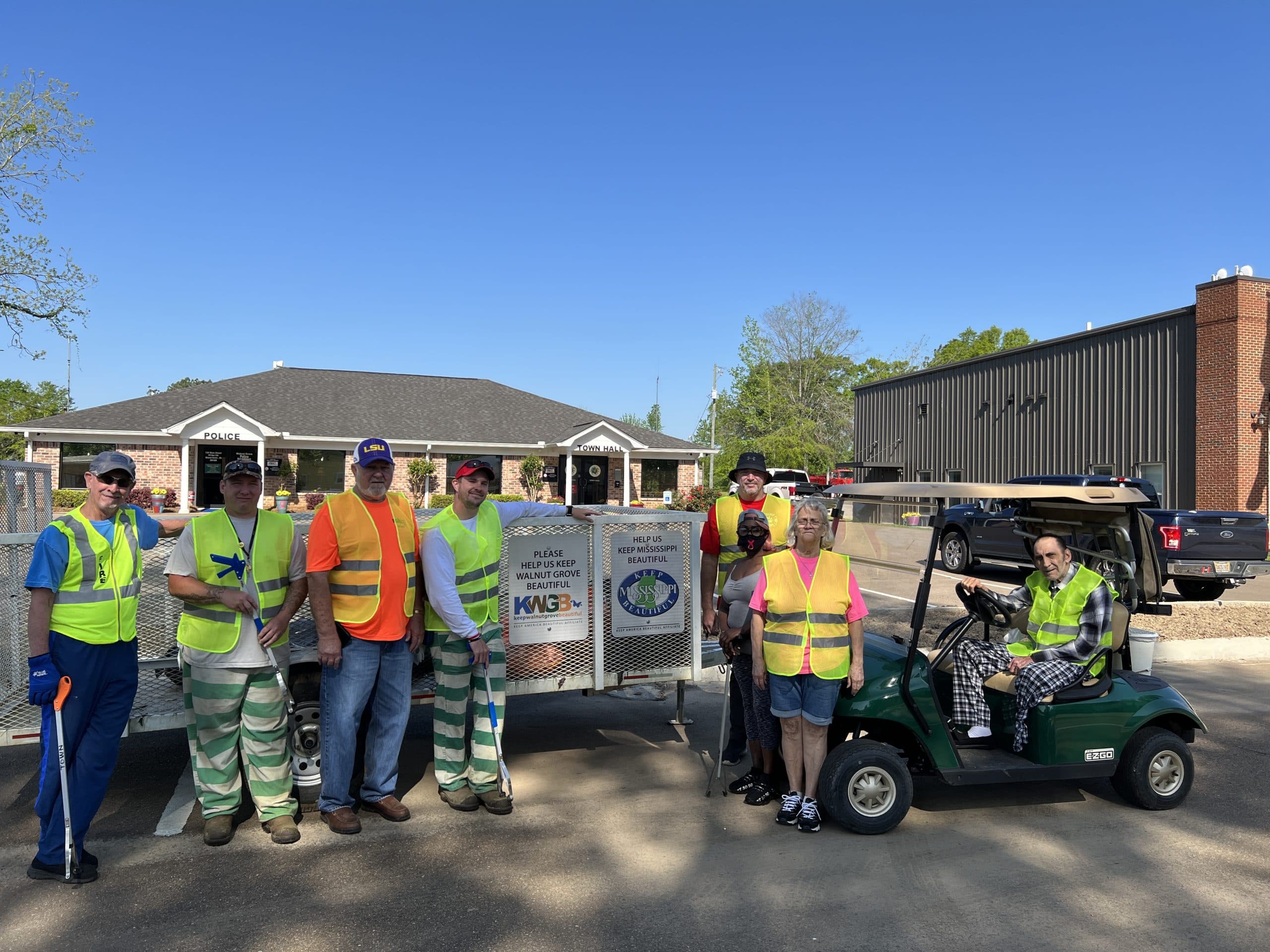 Crew in yellow and orange vests by a trailer outside a brick town hall, a sign reading "Please help us keep Walnut Grove beautiful"; a man sits in a green golf cart