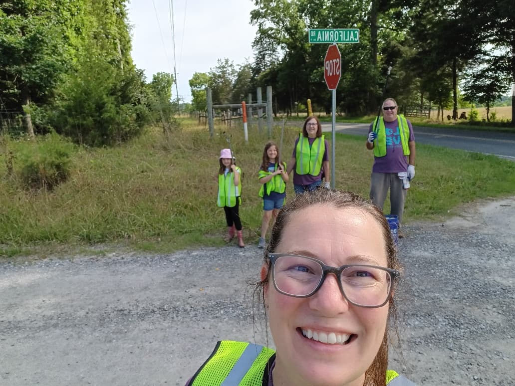 Selfie of a woman in a neon vest on a gravel roadside, with two girls, another woman and a man in vests behind her under a "California Rd" street sign and stop sign