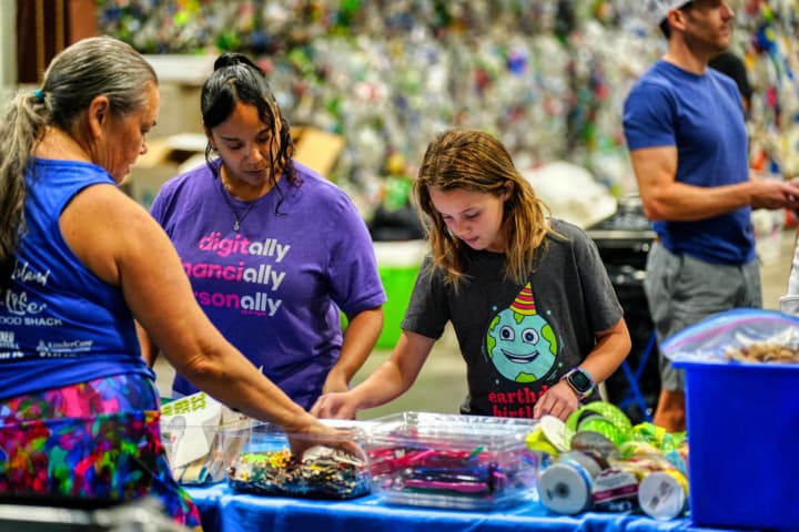 Three people sort small collected items on a blue-draped table inside a recycling facility, bales of crushed plastic bottles stacked behind them