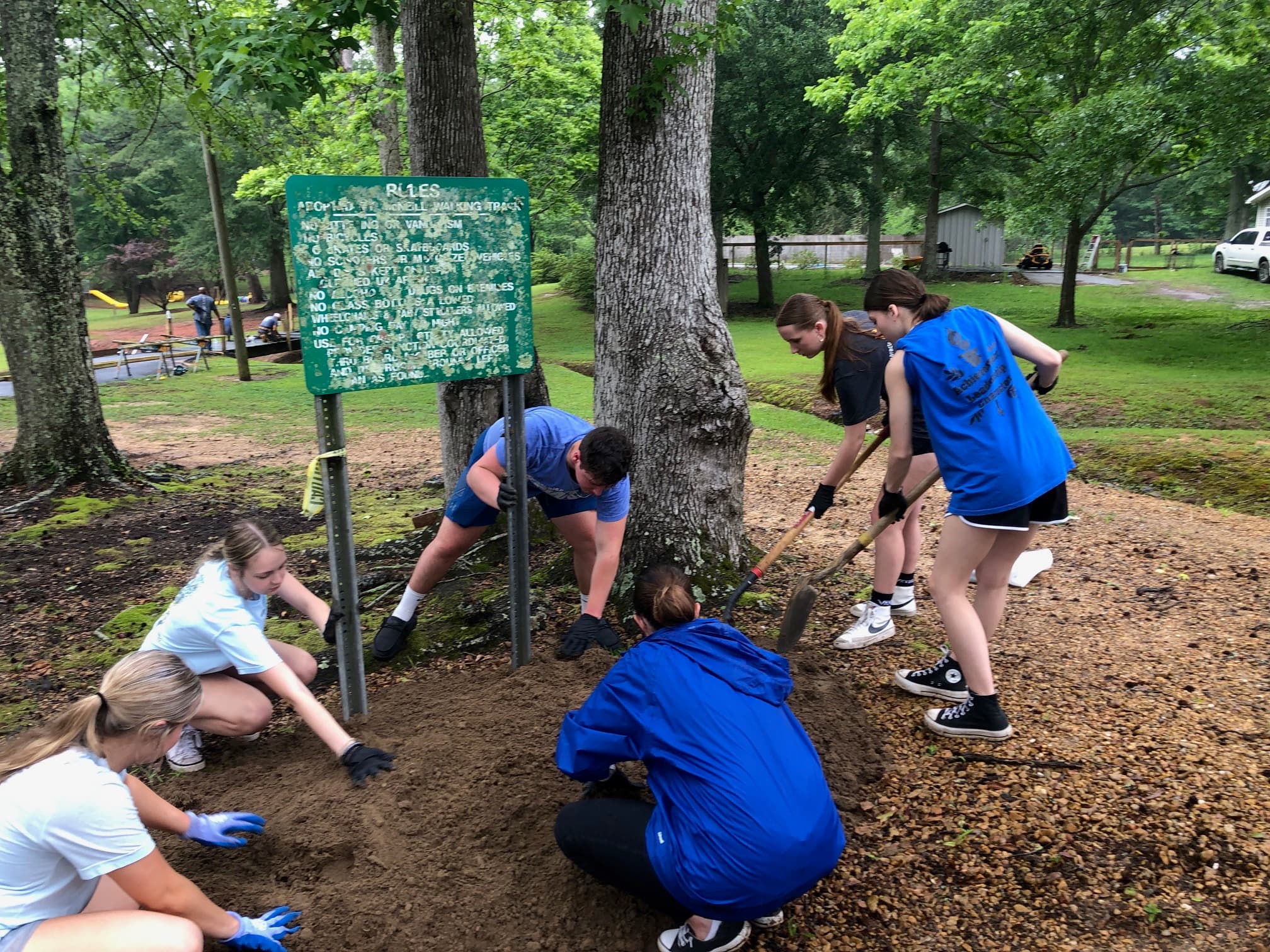 Teenagers in blue and white shirts spread and rake fresh gravel around the base of a weathered green park "Rules" sign along a shaded walking track
