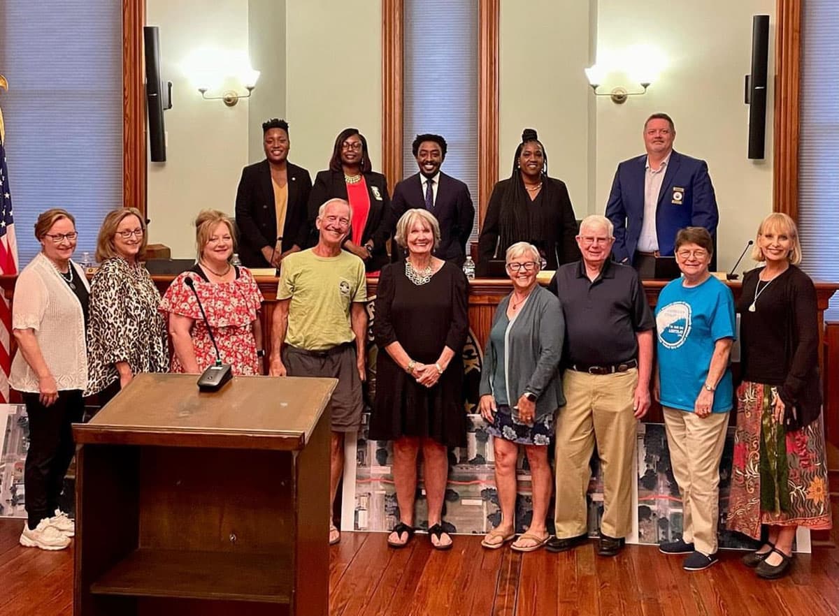 Fifteen people pose in two rows in a wood-panelled council chamber, five officials standing behind the dais and residents in front of a wooden lectern