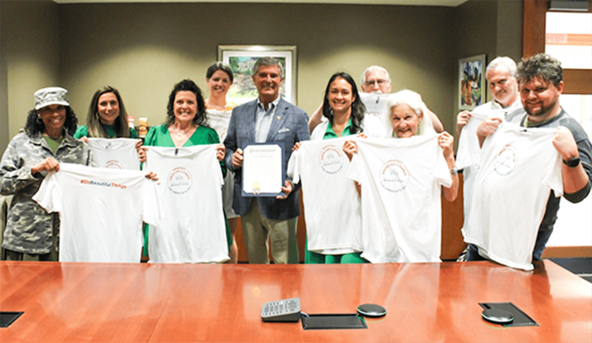 Ten people around a boardroom table hold up white T-shirts, one reading "#DoBeautifulThings", while a man in a checked blazer displays a framed proclamation