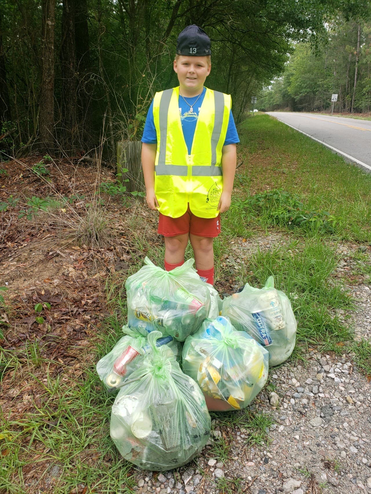 Boy in a neon safety vest and red shorts stands on a rural roadside behind five clear green bags packed with collected cans and bottles