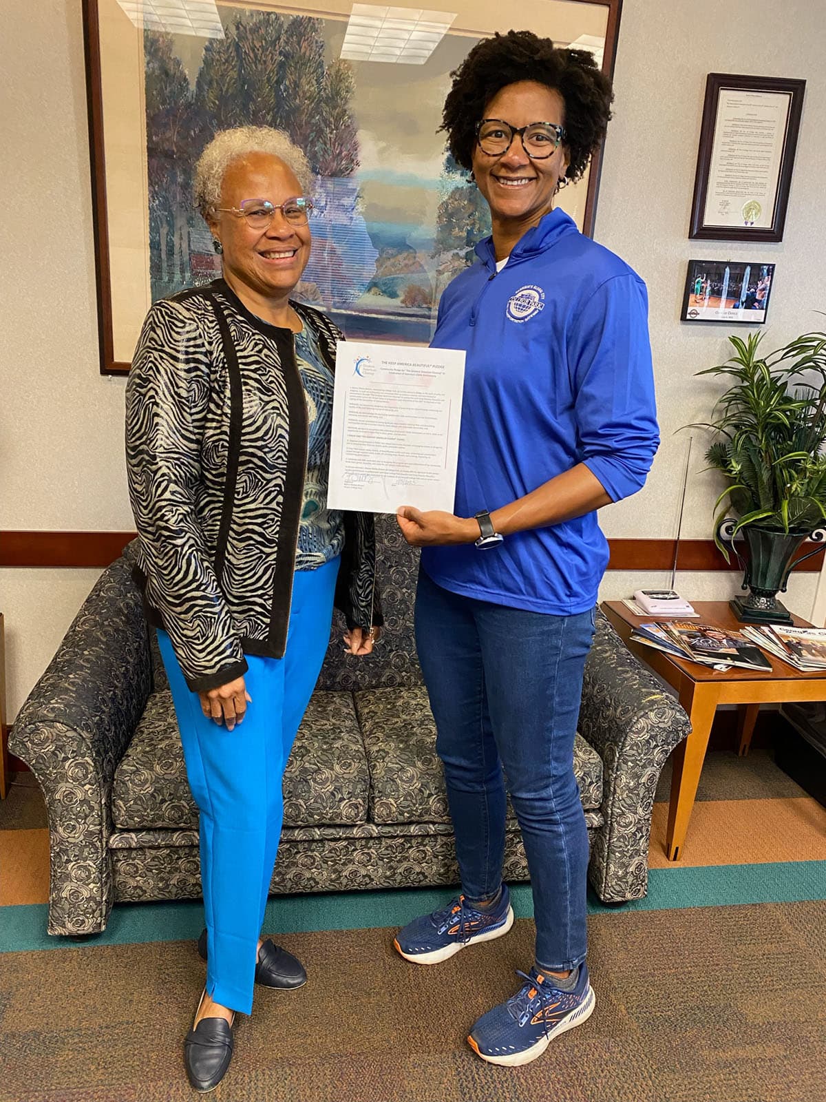 Two women stand in a carpeted office holding a signed sheet headed "The Keep America Beautiful Pledge", a framed landscape painting behind them