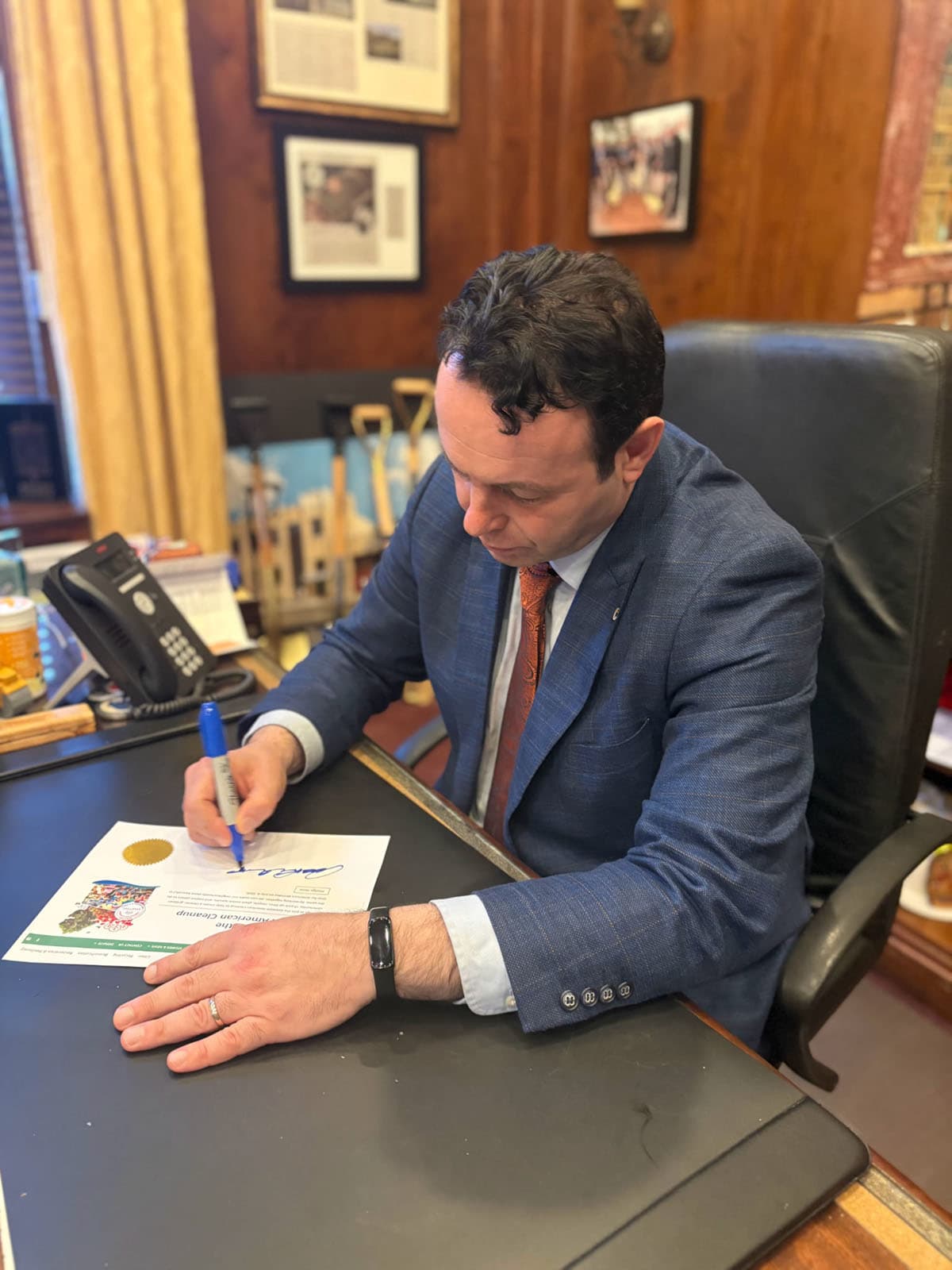 Man in a blue checked suit signs a Great American Cleanup pledge with a marker at a wood-panelled office desk lined with ceremonial shovels