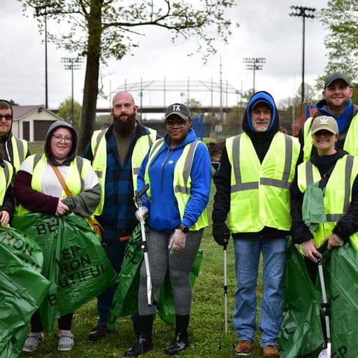 Eight volunteers in neon vests and winter layers stand on a park lawn holding green cleanup bags, stadium light poles and a ballfield behind them