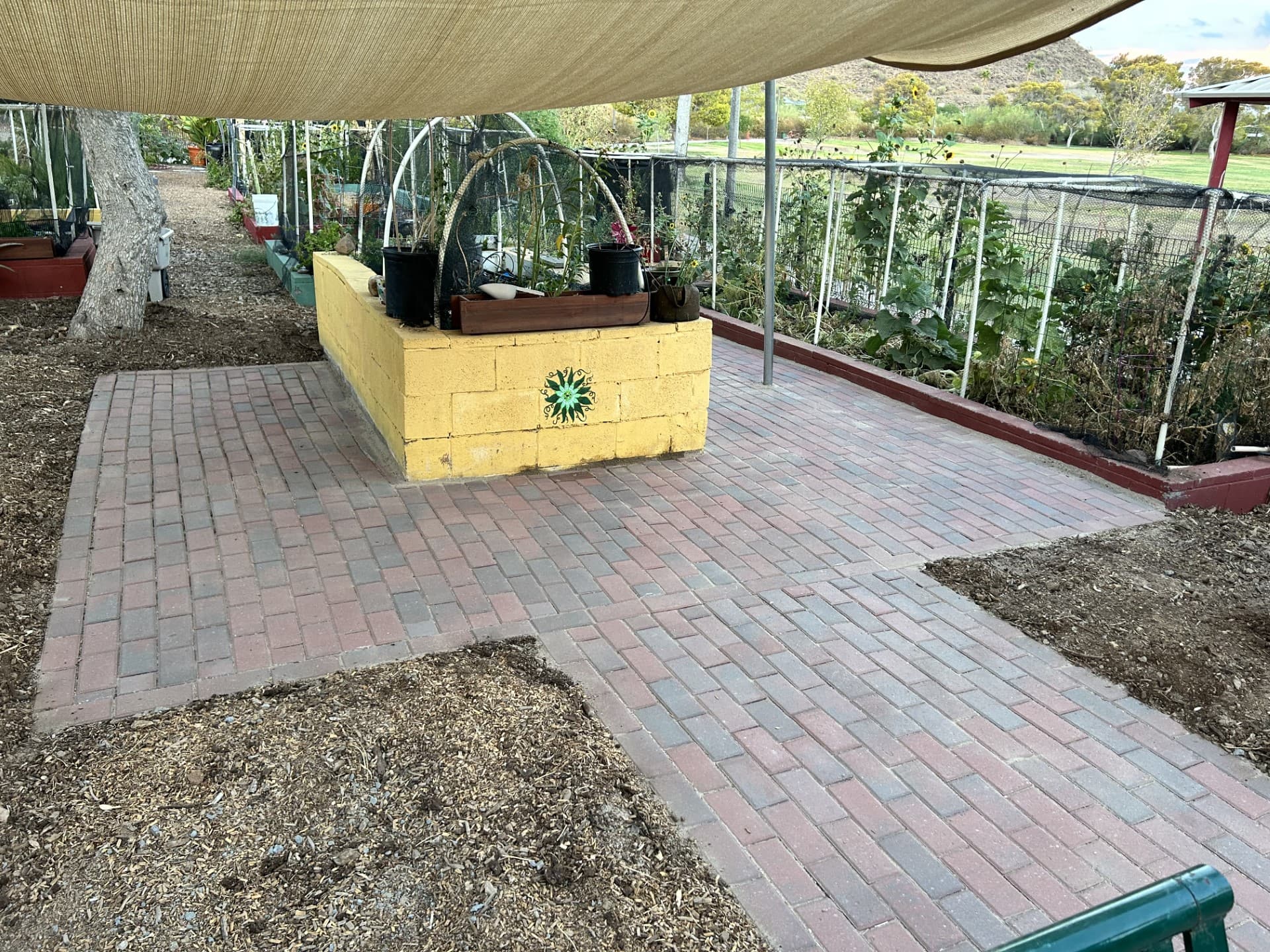 Newly laid brick paver patio in a community garden beneath a tan shade sail, beside a yellow block planter wall and fenced vegetable beds