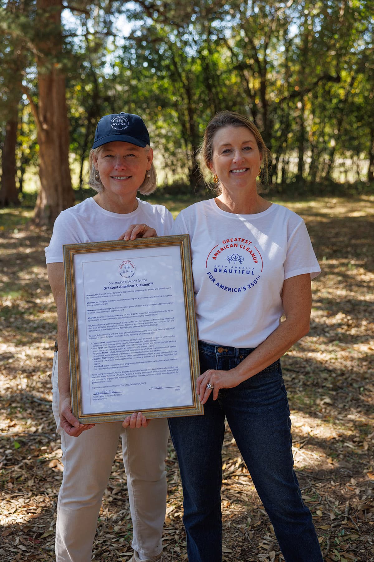 Two women stand in dappled woodland holding a gold-framed document headed Declaration of Action for the Greatest American Cleanup. The left wears a navy cap and white tee; the right wears a white tee printed GREATEST AMERICAN CLEANUP, KEEP AMERICA BEAUTIFUL, FOR AMERICA'S 250TH.